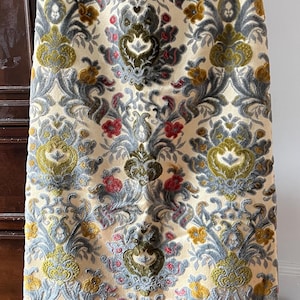 May include: A long, cream-colored skirt with an ornate floral pattern. The design features repeating motifs in shades of green, gray, red, and yellow. The skirt is made of a soft, velvety fabric, and is hanging on a hanger.