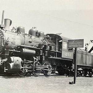 May include: Black and white photograph of a steam locomotive, number 211, with a sign that reads "BE SURE ENGINE'S UNTIL CYLINDERS ARE FULL FRESH WATER". The locomotive is parked near a water tower.