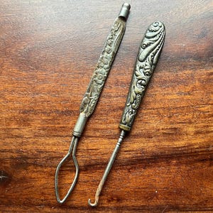 May include: Two antique silver tools with ornate floral and scroll designs. One tool has a loop at the end, and the other has a hook. The tools are set against a warm, brown wooden surface.