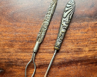 May include: Two antique silver tools with ornate floral and scroll designs. One tool has a loop at the end, and the other has a hook. The tools are set against a warm, brown wooden surface.