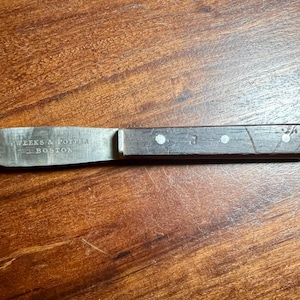 May include: A vintage butter knife with a wooden handle and a silver blade. The blade is stamped with "WEEKS & POTTER BOSTON". The handle is dark brown with three visible rivets. The knife is resting on a wooden surface.