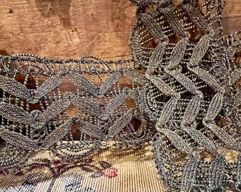 May include: Close-up of decorative silver and black lace trim with a leaf-like pattern. The trim is placed on a patterned fabric with floral elements, creating a vintage aesthetic. The intricate design and metallic threads add a touch of elegance.
