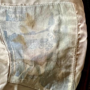 May include: Close-up of the inside of a garment, showing a light tan lining with a rectangular pocket. The pocket features a faded, patterned design in shades of blue, gray, and brown. The stitching is visible, and the overall texture appears soft.