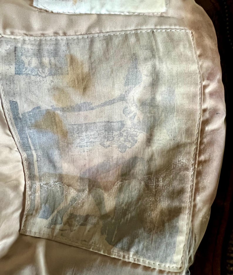 May include: Close-up of the inside of a garment, showing a light tan lining with a rectangular pocket. The pocket features a faded, patterned design in shades of blue, gray, and brown. The stitching is visible, and the overall texture appears soft.