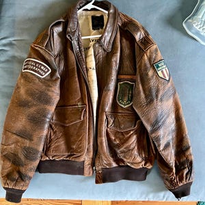 May include: A brown leather bomber jacket with a distressed finish. Features include a zip closure, multiple pockets, and patches that read "OFFICIAL US WAR PHOTOGRAPHER" and "WAR CORRESPONDENT EUROPE". The jacket has a ribbed hem and cuffs.