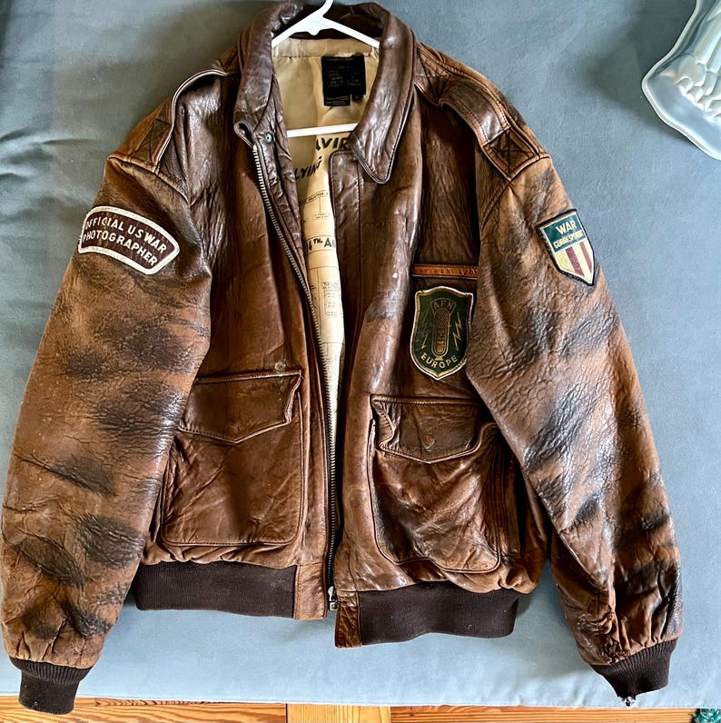 May include: A brown leather bomber jacket with a distressed finish. Features include a zip closure, multiple pockets, and patches that read "OFFICIAL US WAR PHOTOGRAPHER" and "WAR CORRESPONDENT EUROPE". The jacket has a ribbed hem and cuffs.