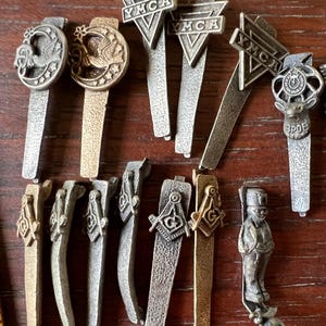 May include: A collection of vintage metal lapel pins and tie clips. Designs include the YMCA logo, Masonic symbols, and other decorative emblems. The pins are in various metallic finishes, including silver and gold, and are arranged on a wooden surface.