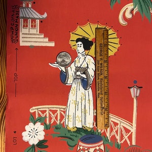May include: A red wallpaper sheet featuring a printed illustration of a figure holding a parasol, a pagoda, and a bridge. A wooden ruler measuring 6 inches is placed vertically next to the figure, and a silver coin rests on the figure's hand.