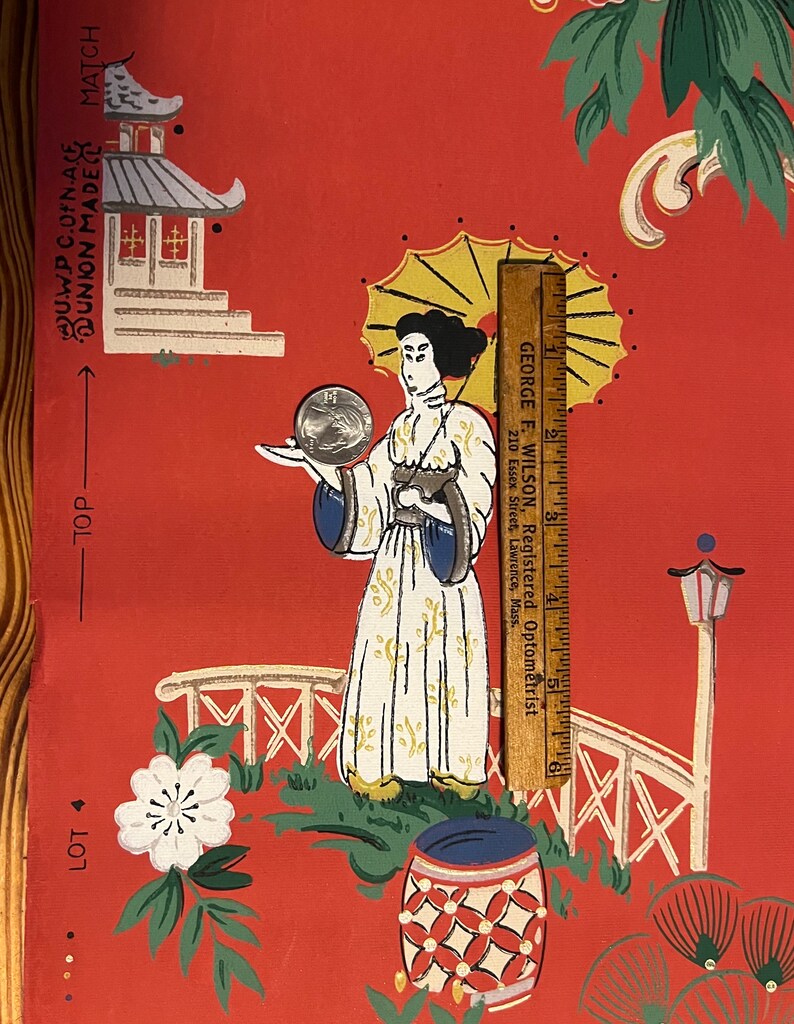 May include: A red wallpaper sheet featuring a printed illustration of a figure holding a parasol, a pagoda, and a bridge. A wooden ruler measuring 6 inches is placed vertically next to the figure, and a silver coin rests on the figure's hand.