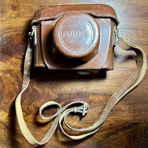 May include: A vintage brown leather camera case with a matching strap. The camera case has a circular lens cover with the word "FUTURA" embossed on it. The strap is attached to the case with metal hardware.