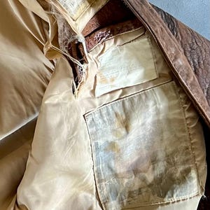 May include: Close-up of the interior of a vintage brown leather jacket. The lining is a light tan color, with visible pockets and labels. The jacket shows signs of wear and tear, with some areas of damage.