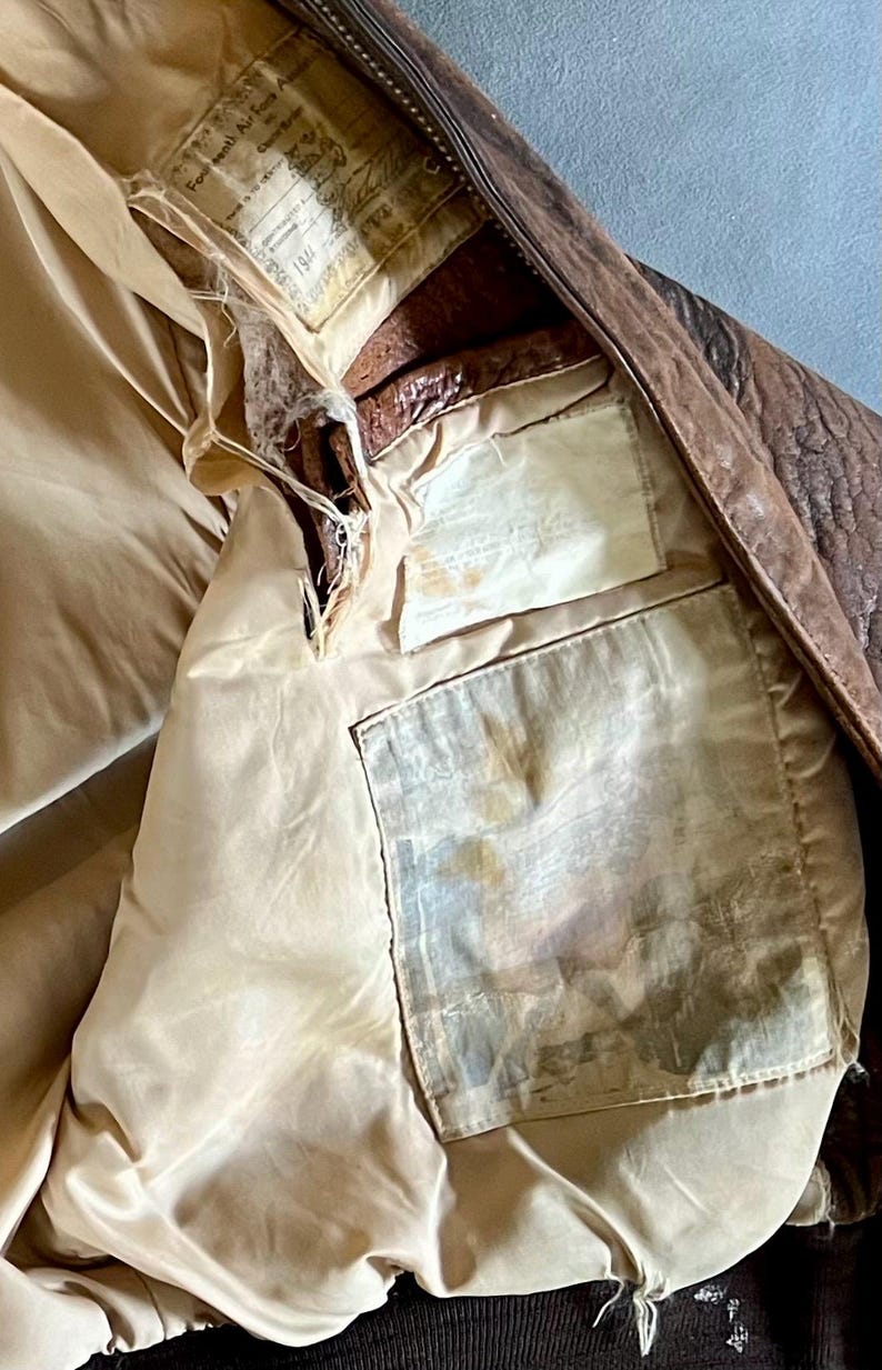 May include: Close-up of the interior of a vintage brown leather jacket. The lining is a light tan color, with visible pockets and labels. The jacket shows signs of wear and tear, with some areas of damage.