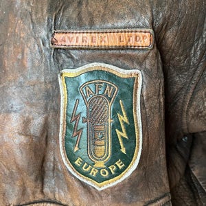 May include: Close-up of a brown leather jacket with a distressed finish. Features a patch with a microphone graphic, lightning bolts, and the text "AFN EUROPE". The jacket also has a small rectangular patch that reads "AVIREX LTD."