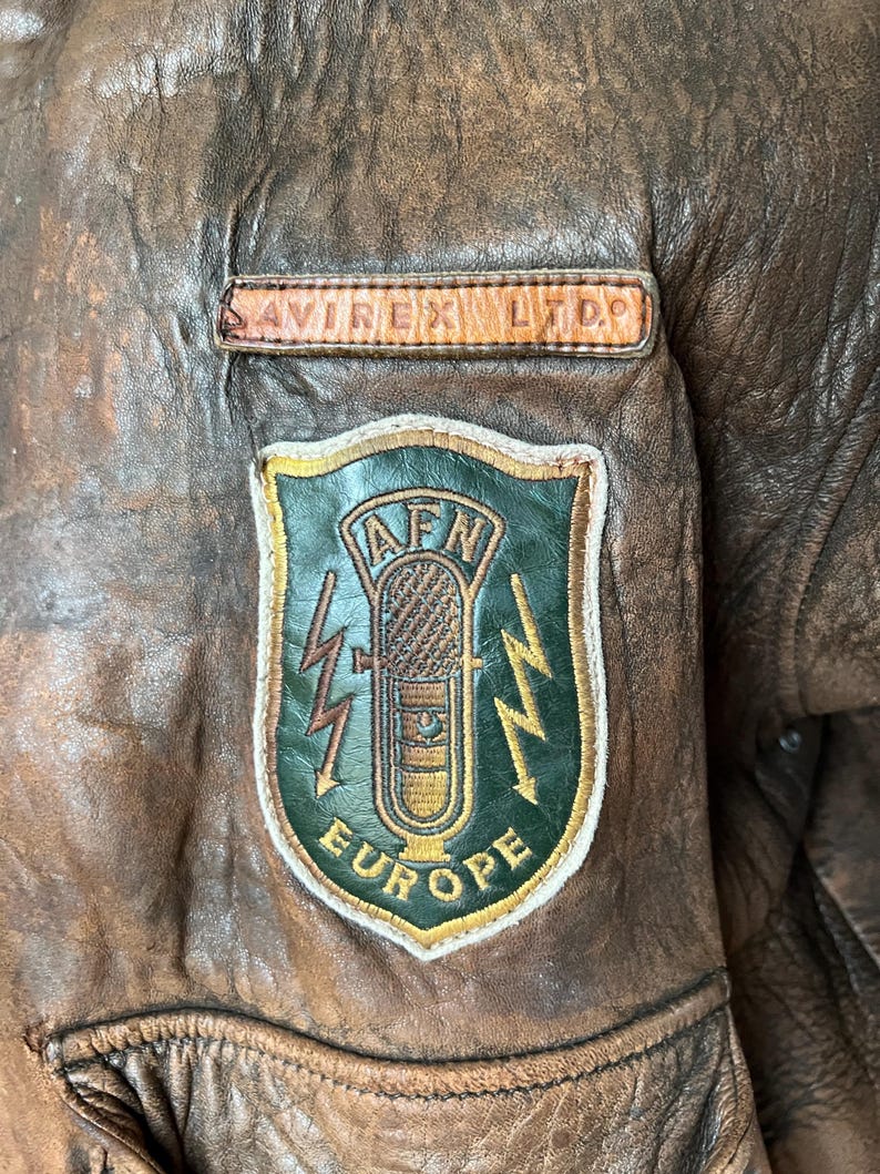 May include: Close-up of a brown leather jacket with a distressed finish. Features a patch with a microphone graphic, lightning bolts, and the text "AFN EUROPE". The jacket also has a small rectangular patch that reads "AVIREX LTD."