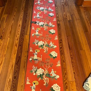 May include: A long roll of wallpaper featuring a vibrant red background with a repeating pattern of figures holding yellow umbrellas, arched bridges, pagodas, and large white and green floral motifs. The paper is unrolled vertically.