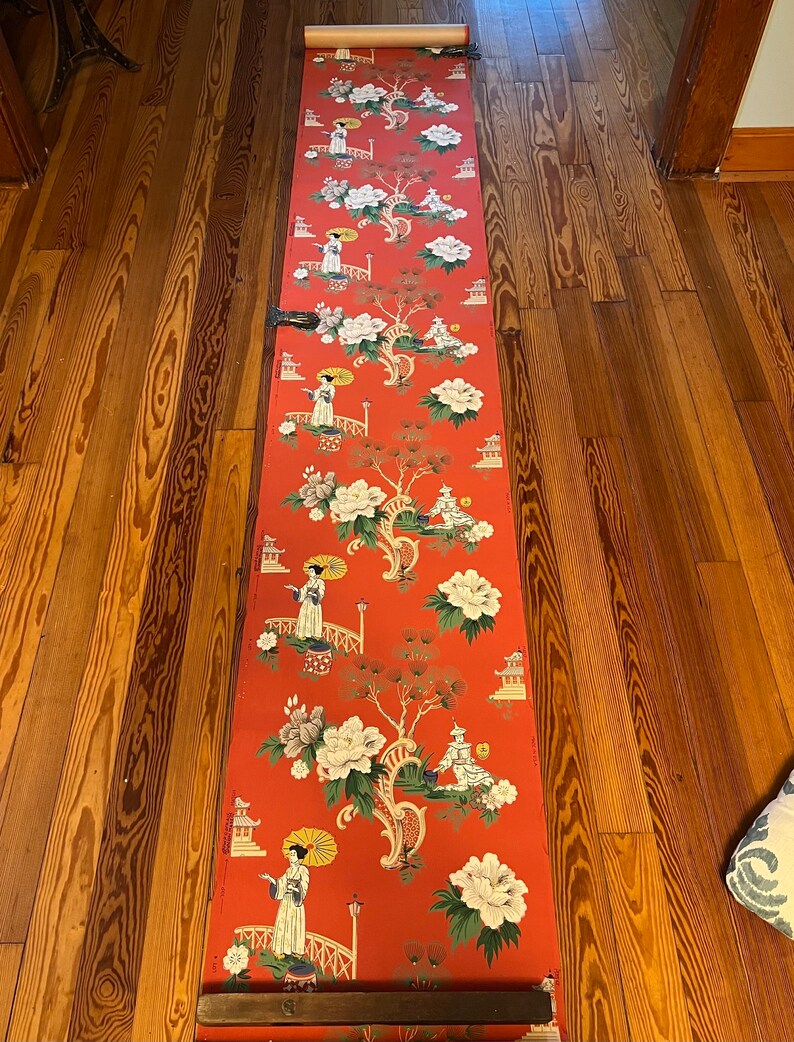 May include: A long roll of wallpaper featuring a vibrant red background with a repeating pattern of figures holding yellow umbrellas, arched bridges, pagodas, and large white and green floral motifs. The paper is unrolled vertically.