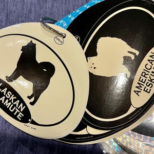 May include: A set of dog breed decals, including an Alaskan Malamute and an American Eskimo, attached with a metal ring. The decals feature black silhouettes of the dogs on a cream background, with text identifying the breeds.