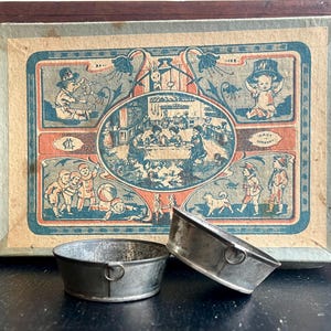 May include: Two small, vintage metal baking molds with a decorative box. The box features a colorful illustration with children and scenes, with the text "MADE IN GERMANY". The molds are round with a small handle.