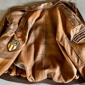 May include: A brown leather jacket with a distressed finish. The jacket features patches that read "WAR CORRESPONDENT" and "OFFICIAL US WAR PHOTOGRAPHER". The jacket has a zipper closure and a ribbed hem.