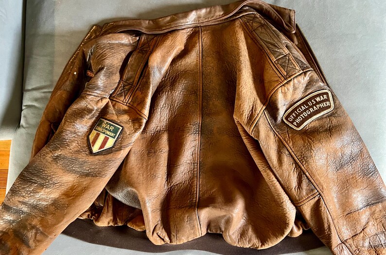 May include: A brown leather jacket with a distressed finish. The jacket features patches that read "WAR CORRESPONDENT" and "OFFICIAL US WAR PHOTOGRAPHER". The jacket has a zipper closure and a ribbed hem.