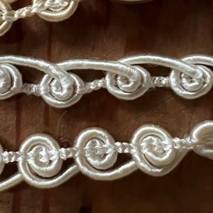 May include: Close-up of decorative trim with a looped design. The trim is a creamy white color and appears to be made of a satin-like material. The loops are evenly spaced and create a repeating pattern. The background is a wooden surface.