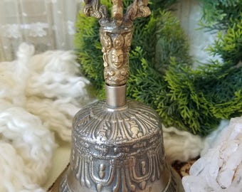May include: A silver-colored bell with ornate detailing and a decorative handle. The handle features a detailed design with a face and a curved top. The bell is likely made of metal and has a vintage or antique appearance.