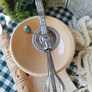 May include: An antique hand-crank egg beater with a clear handle and a cream-colored mixing bowl. The beater is made of metal and has a green handle. A wooden rolling pin with carved designs is also visible.