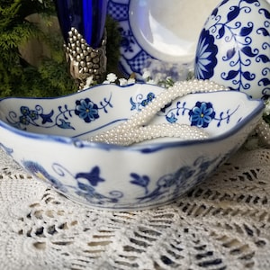May include: A white porcelain bowl with a scalloped edge, adorned with blue floral patterns. A strand of white pearls rests inside the bowl. The bowl is part of a collection of blue and white decorative items.