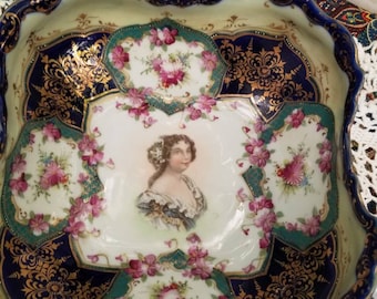 May include: A large, scalloped porcelain bowl with a blue and gold rim. The bowl is decorated with a floral pattern and a portrait of a woman in the center. The bowl is perfect for serving fruit, snacks, or other treats.