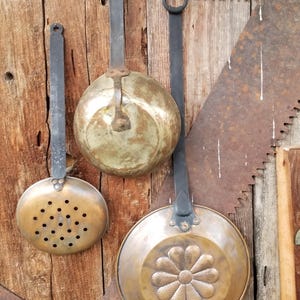 May include: Three vintage metal kitchen tools with long dark iron handles. One is a round brass skimmer with perforated holes, one is a smooth brass bowl, and one is a brass pan featuring a central embossed flower design on the base.