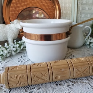 May include: A white ceramic pot with a polished copper band around the center and a long copper handle, positioned alongside a light-colored wooden rolling pin featuring carved geometric patterns and star designs across its cylindrical surface.