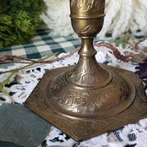 May include: A vintage brass candlestick holder with an ornate, floral design. The base is square with rounded corners and detailed leaf patterns. The candlestick is a warm gold color, adding a touch of antique charm to any decor.