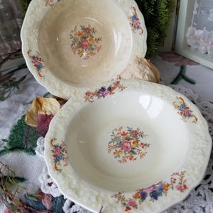 May include: Two cream-colored, scalloped-edge bowls with floral designs. Each bowl features a central floral bouquet and smaller floral clusters around the rim. The bowls are vintage and have a slightly aged appearance.