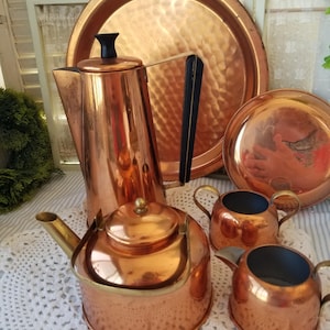 May include: A collection of copper kitchenware, including a tall coffee pot with a black handle and lid, a kettle, two creamers, and a large serving tray. The items have a hammered texture and a shiny, reflective surface.