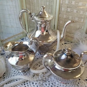 May include: A silver tea set with a teapot, creamer, and sugar bowl. The set is on a white crocheted doily with a pearl necklace draped over it.