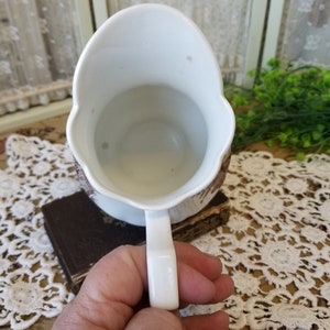May include: White ceramic pitcher with a brown and white floral design. The pitcher has a handle and a wide mouth.