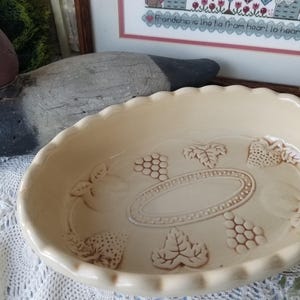 May include: Cream-colored ceramic pie dish with a scalloped edge. The dish features embossed grape and leaf designs around the rim and center. A decorative kitchenware item.