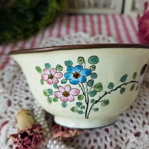 May include: A small, white ceramic bowl with a brown rim. The bowl is decorated with a floral design featuring pink, blue, and green flowers.