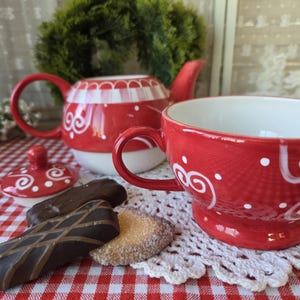 May include: A red ceramic teapot and matching teacup featuring white hand-painted swirl and dot patterns. The teapot has a white lid with red swirls, and the set is accompanied by two dark chocolate-covered cookies and a round sugar cookie.