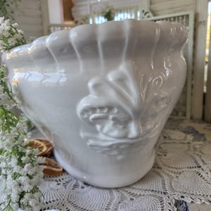 May include: A glossy white ceramic planter featuring a scalloped rim and a prominent embossed relief design of a face on the front. The bowl-shaped vessel has a smooth, reflective finish and is decorated with small white flowers along the side.