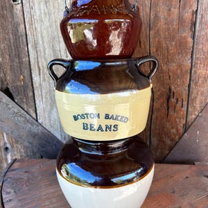 May include: A stack of three ceramic bean pots. The top pot is dark brown with a wire handle and the word BEANS. The middle pot is two-toned brown and tan with two handles and the text BOSTON BAKED BEANS. The bottom pot is brown and cream.
