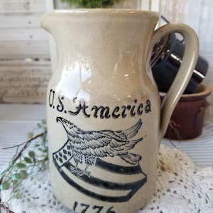 May include: A beige stoneware pitcher with a black and white design of an eagle and the words "U.S. America 1776".