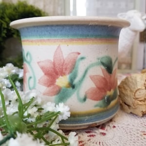 May include: A ceramic planter with a floral design. The planter features a cream-colored base with pink flowers and green stems. The rim has a blue, yellow, and pink band. The planter is suitable for small plants and adds a touch of vintage charm.