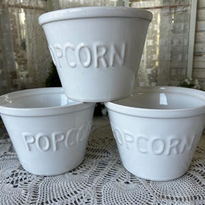 May include: Three white ceramic bowls with a tapered shape and a wide rim. Each bowl features the word POPCORN embossed in capital letters on the front. The bowls are arranged in a stack with one bowl placed on top of two others.