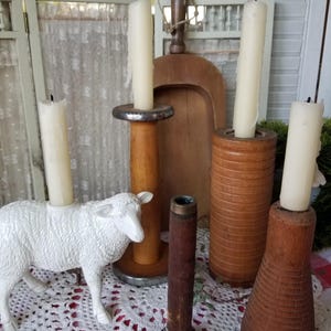 May include: A collection of vintage candle holders and a white sheep figurine on a white lace tablecloth. The candle holders are made of wood and metal, and hold white candles. A wooden scoop and a wooden spool are also present.