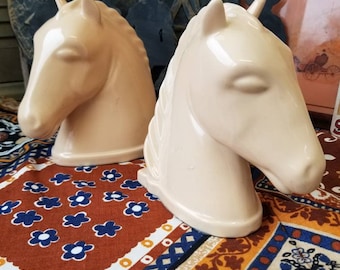 May include: Two ceramic horse head bookends in a pale cream color. The bookends are sitting on a colorful patterned fabric.