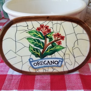 May include: Oval ceramic serving dish with a brown rim, featuring a hand-painted oregano design. The design includes green leaves, red flowers, and the word "OREGANO" on a blue banner. The background has a cracked tile pattern.