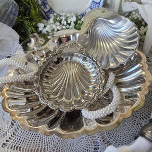 May include: A set of three silver-toned metal dishes shaped like scallop shells with fluted edges. The dishes are stacked, featuring a polished metallic finish. A strand of white faux pearls is draped over the shells, adding texture to the display.