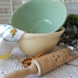 May include: A light green ceramic mixing bowl sits atop a cream-colored bowl. A wooden rolling pin with carved floral designs and a metal whisk rest on a white lace tablecloth. An embroidered cloth adds a touch of color.