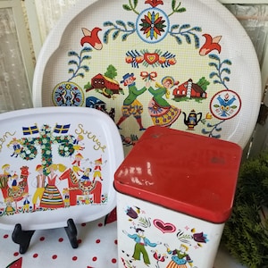May include: Two white metal trays with a Swedish folk art design. One tray features a maypole and dancing figures. The other tray features a couple dancing and a red barn. A red and white tin box with a heart and a couple dancing. The text on the tin box reads "Arden Finest Hard Candies Filled x Solid".